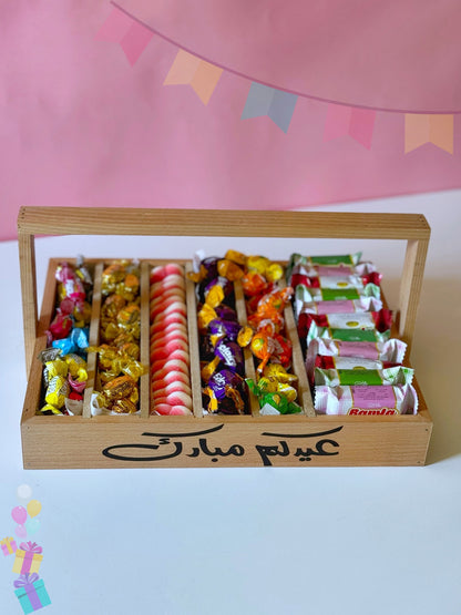 A wooden Eid sweets tray engraved with "Eidkum Mubarak," filled with an assortment of bonbons, jellies, and biscuits, placed on a white surface.