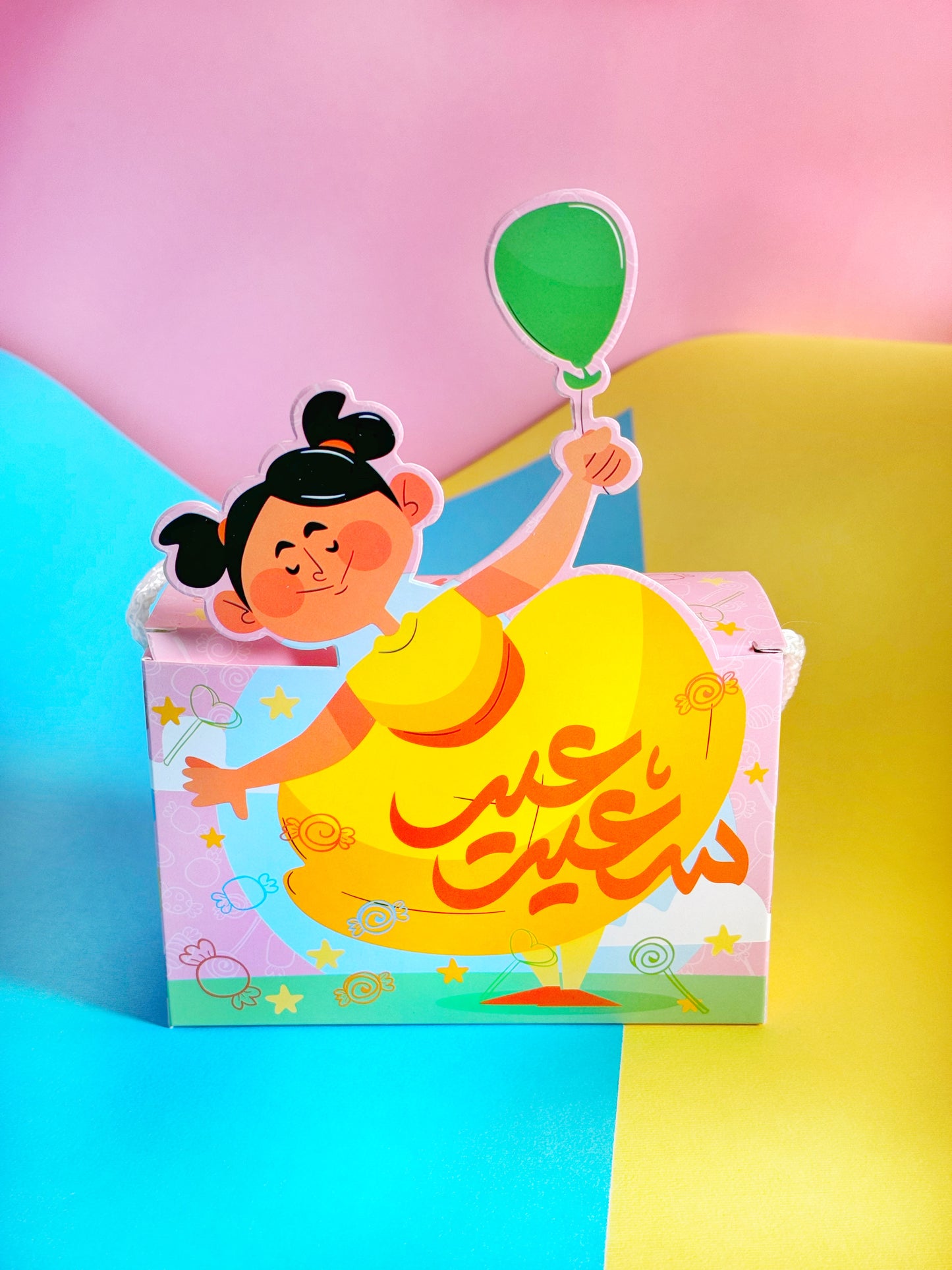 A vibrant Eid Joy Bag in a girl variant, featuring an assortment of sweets and a fun water game toy, set against a pink and yellow background.