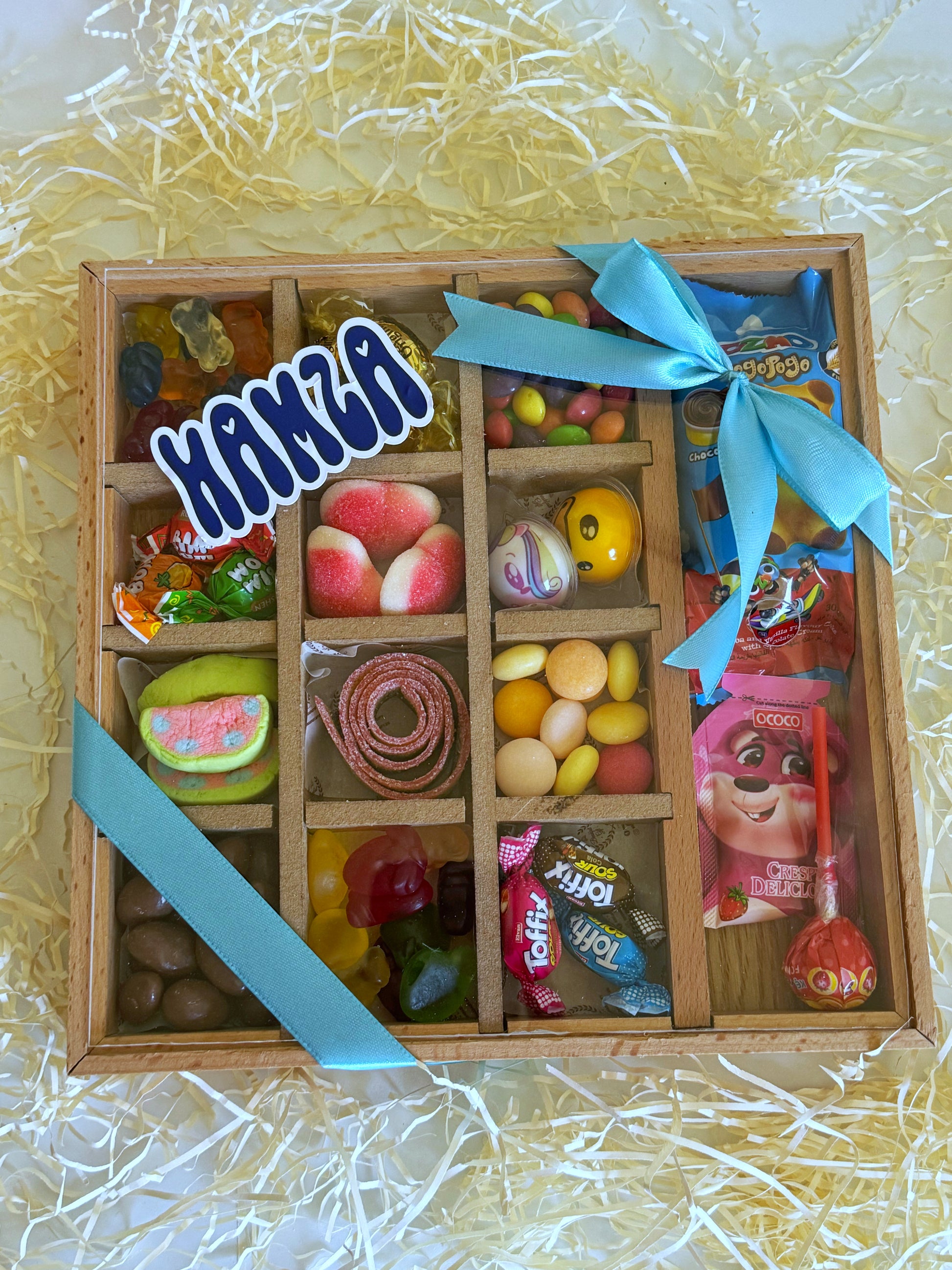 A beautifully arranged Eid gift featuring a wooden divided box filled with colorful candies, chocolates, and festive treats, wrapped with a blue ribbon.