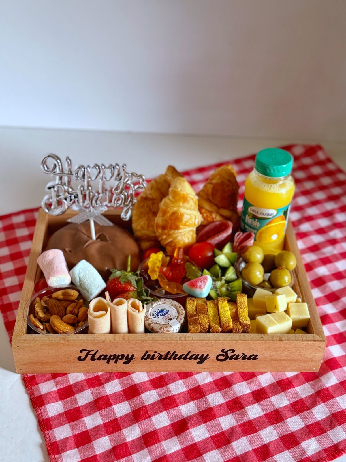 A beautifully crafted wooden tray with "Happy birthday Sara" message, featuring croissants, cheddar cheese, beef salami, strawberries, kiwi, cucumbers, cherry tomatoes, Turkish delights, jam, Marshmallows, candy, nuts, olives, and a bottle of fresh juice. Perfect for a joyful breakfast or special occasion.