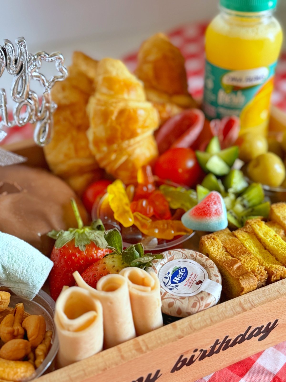 Close-up of the Happiness in a Tray showcasing croissants, cheddar cheese, Turkish delights, candy, nuts, olives, and a fresh juice bottle. The tray is personalized with a "Happy birthday Sara" message, ideal for a festive celebration.