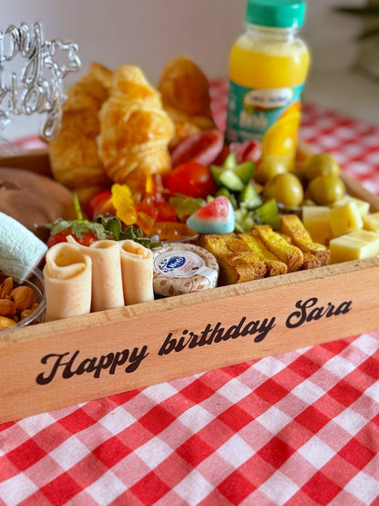 A vibrant assortment of pastries, fruits, nuts, and cheese on a personalized wooden tray with "Happy birthday Sara" text. Includes croissants, cheddar cheese, Turkish delights, candy, olives, and fresh juice for a cheerful breakfast experience.