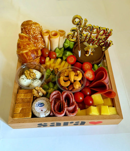 A wooden tray labeled "Sara ❤️" filled with croissant, personalized glass, cake, cheeses, fruits, cold cuts, and nuts.