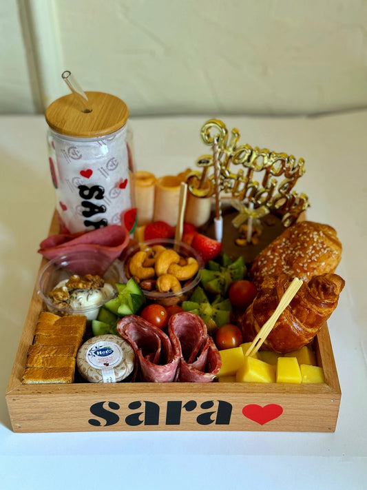 A wooden tray labeled "Sara ❤️" filled with croissant, personalized glass, cake, cheeses, fruits, cold cuts, and nuts.

