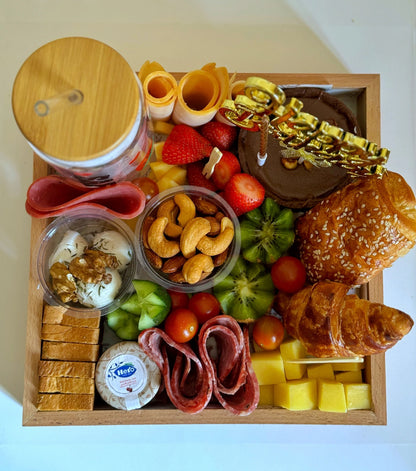 Top-down view of a personalized birthday breakfast tray with mini chocolate cake, fruits, turkey rolls, and mixed snacks.