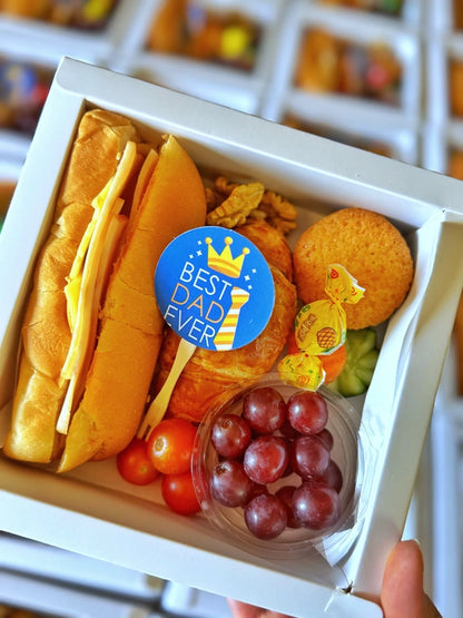 A colorful open Little Cheers Box showing a mini sandwich, muffin, croissant, fresh fruits, veggies, candy, and nuts, all neatly arranged in an 18×18 cm tray.