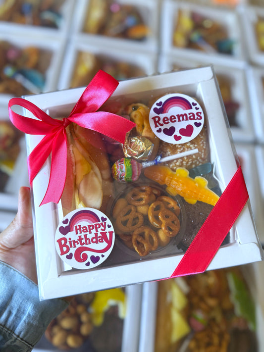 A hand holding a closed Mini Bites Birthday Box with a red ribbon and “Happy Birthday” sticker, showcasing its cheerful packaging.