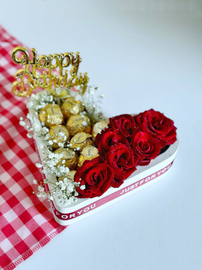 A heart-shaped box filled with Fantancy Rocher-style Milk and Nut Chocolates and vibrant red roses, decorated with delicate white baby's breath flowers and a golden "Happy Birthday" topper on a red checkered cloth.