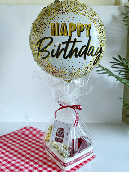 A beautifully wrapped Roses & Chocolate Heart gift box with a "Happy Birthday" balloon and "Just for You" ribbon, elegantly arranged on a red checkered cloth, ready for gifting.