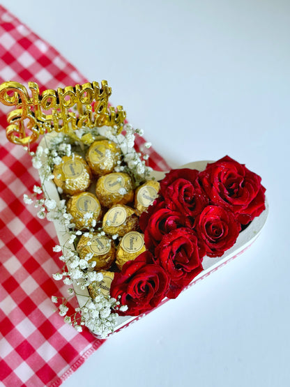 A close-up of a heart-shaped box filled with red roses and Fantancy Rocher-style chocolates, accented with white baby's breath flowers and wrapped with a "Just for You" ribbon on a red checkered cloth.