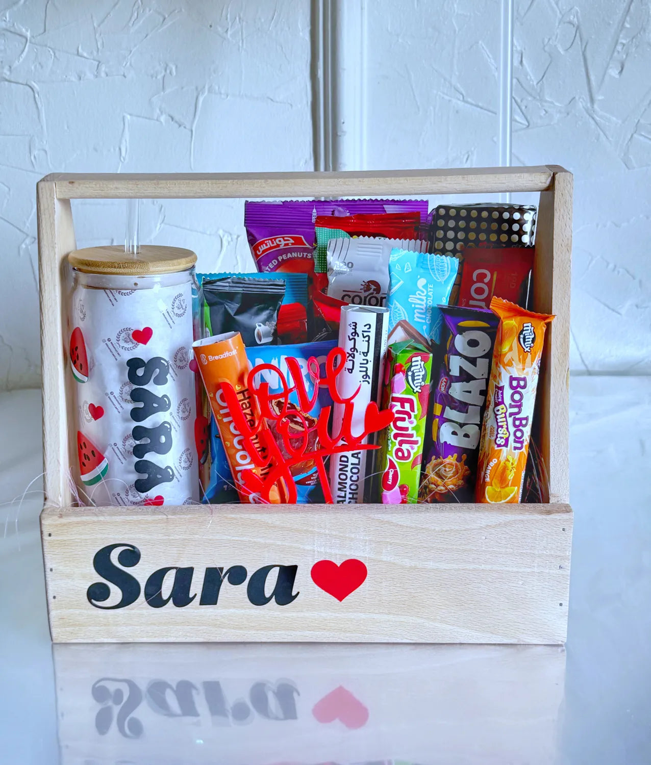A wooden gift crate labeled "Sara ❤️" filled with chocolates, candy, snacks, and a personalized iced coffee glass decorated with watermelon slices and red hearts.