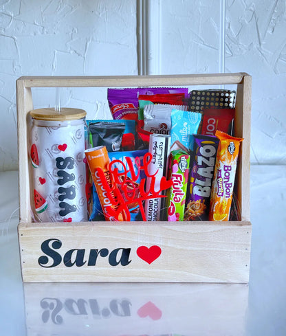 A wooden gift crate labeled "Sara ❤️" filled with chocolates, candy, snacks, and a personalized iced coffee glass decorated with watermelon slices and red hearts.