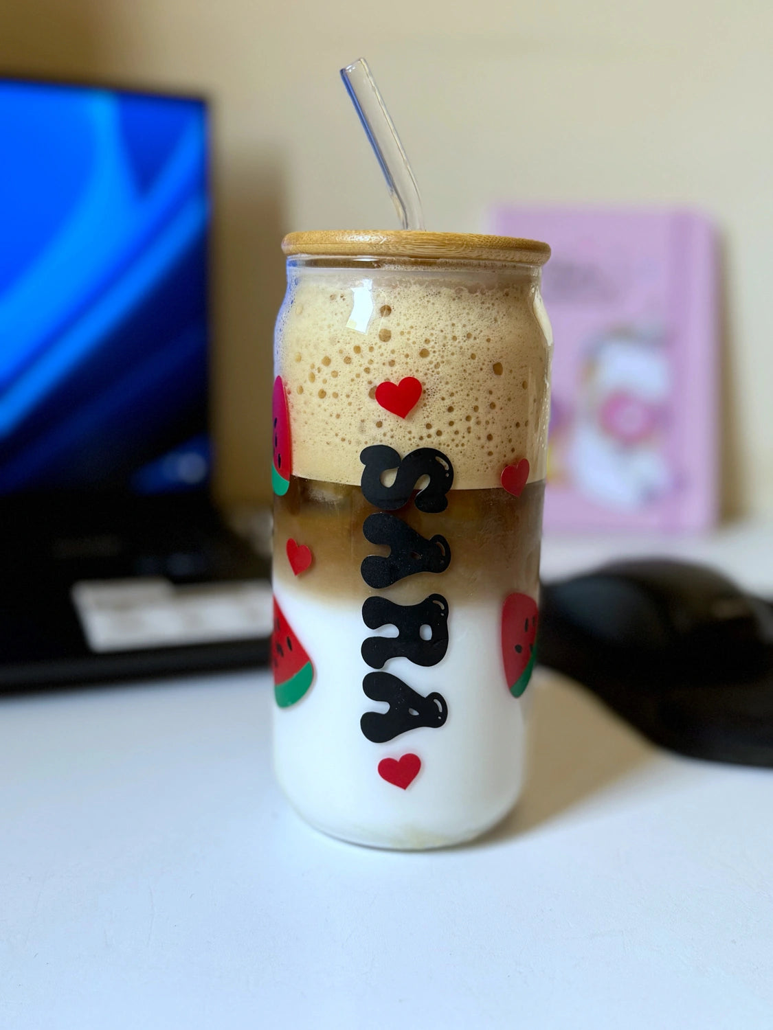 Close-up of a customized glass iced coffee cup with “Sara” written in bold letters, red hearts, and watermelon designs, sitting on a desk.