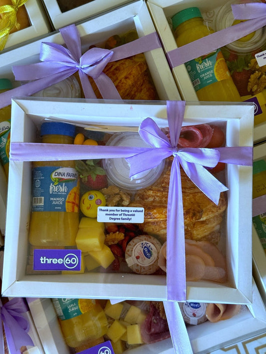 A corporate breakfast box with a purple ribbon, featuring croissant, juice, cheese cubes, cherry tomatoes, salami, turkey slices, and a custom Three60 label.