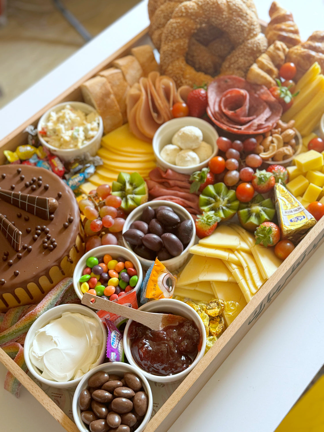 A large wooden tray filled with chocolate cake, cheeses, cold cuts, fruits, and assorted sweets, arranged beautifully for a celebration.