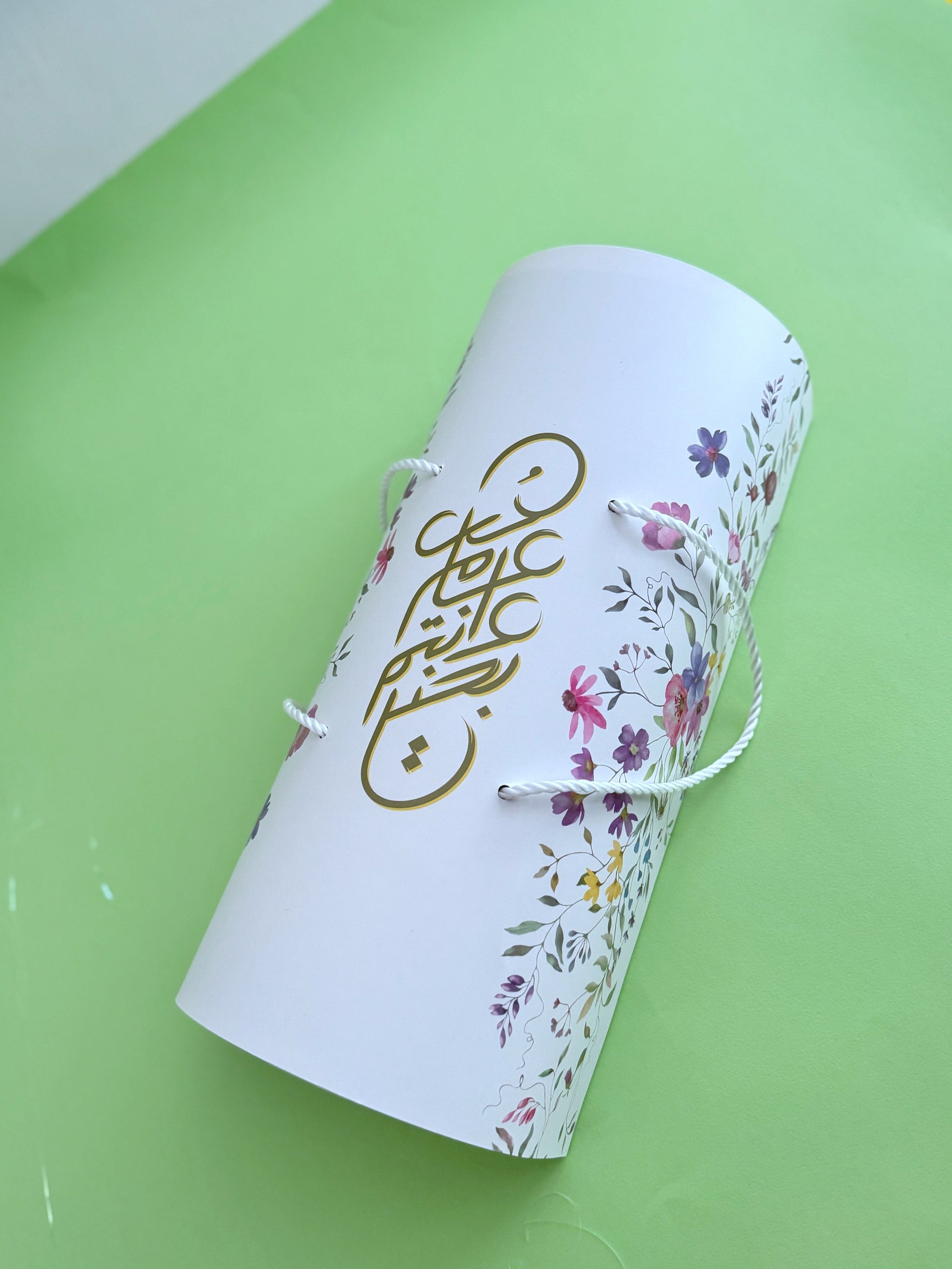 The Grand Kahk Box – Closed floral cylinder gift box, top view showing gold Arabic calligraphy and rope handle